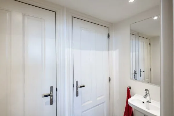 Wit betegelde badkamer met twee witte deuren, een spiegel en een wit fonteintje met rode handdoek.